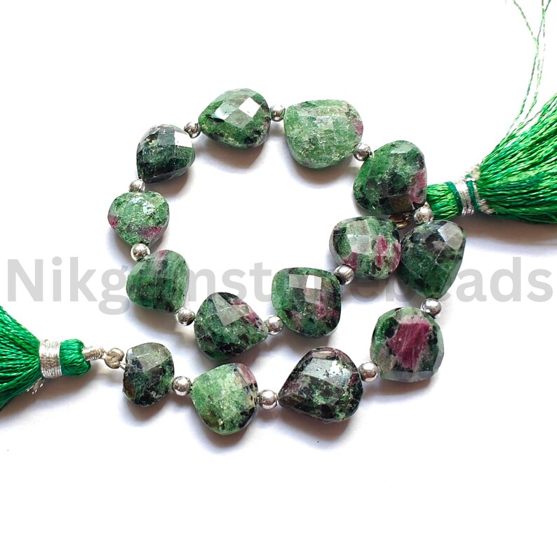 Może przedstawiać: Szczeg&oacute;łowe ujęcie fasetowanych zielonych i czerwonych koralik&oacute;w z kamieni szlachetnych ze srebrnymi przekładkami. Koraliki są nawleczone, tworząc zakrzywiony kształt. Zielone frędzle przymocowane na każdym końcu. Naturalny wygląd.