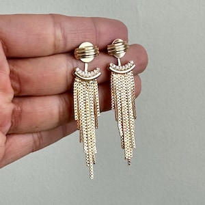 Może przedstawiać: Para złotych kolczyków wiszących z frędzlami z łańcucha i przezroczystymi akcentami z kryształów.