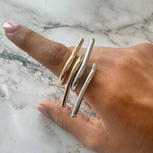 Puede incluir: Tres anillos de oro y plata apilados en un dedo. Los anillos están hechos de metal delgado y curvo y están diseñados para ser usados en el dedo medio.