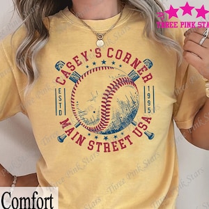 May include: A yellow t-shirt with a baseball graphic and the text "Casey's Corner Est. 1995 Main Street USA".