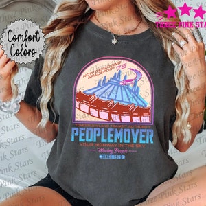 May include: A dark gray t-shirt with a graphic of the Tomorrowland Transit Authority Peoplemover ride at a theme park. The graphic includes the text "Now Departing Spaceport 75" and "Tomorrowland Transit Authority Peoplemover Your Highway in the Sky - Moving People - Since 1975".