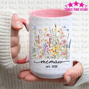 Könnte beinhalten: Weißer Keramikbecher mit rosa Innenraum und farbenfrohem Blumendesign. Der Becher trägt den Schriftzug "memaw" in Schreibschrift und "est. 2025". Der Becher wird von einer Person gehalten.