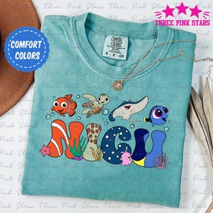 May include: A teal blue t-shirt with a graphic of the characters from the movie Finding Nemo. The characters are arranged to spell out the name "NIGEL". The shirt has a distressed look and is made of a soft, comfortable fabric.