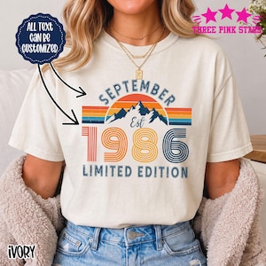 Puede incluir: Una camiseta color marfil con un gráfico de un atardecer, montañas y el texto "SEPTEMBER EST 1986 LIMITED EDITION" en tipografía retro. Una insignia circular azul oscuro en la parte superior izquierda dice "ALL TEXT CAN BE CUSTOMIZED."