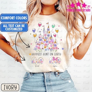 Puede incluir: Camiseta color marfil con un diseño floral de castillo, orejas de Mickey Mouse y un lazo. El texto dice "Happiest Aunt on Earth". La camiseta también tiene el texto "Comfort Colors" y "All text can be customized."