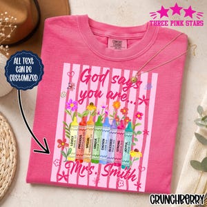 May include: A pink t-shirt with a graphic featuring crayons and the words "God says you are..." and "Mrs. Smith." The crayons are labeled with positive attributes. The shirt also has the text "All text can be customized."