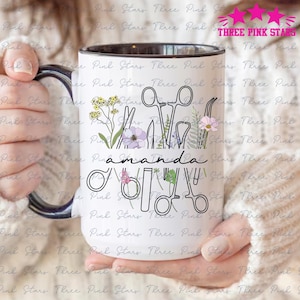 Könnte beinhalten: Eine weiße Keramiktasse mit schwarzem Rand und Henkel. Die Tasse zeigt eine Schwarzweißillustration von chirurgischen Instrumenten und Blumen. Der Text "amanda" ist in schwarzer Kursivschrift geschrieben.