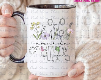 May include: White ceramic mug with a black rim and handle. The mug features a black and white illustration of surgical tools and flowers. The text "amanda" is written in black cursive lettering.