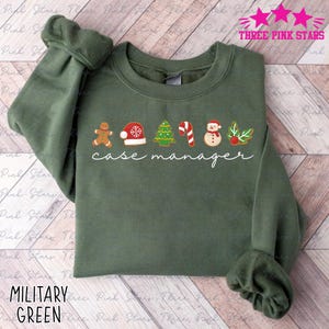 Könnte beinhalten: Militärgrünes Sweatshirt mit dem Schriftzug "case manager" in weißer Schrift. Das Design umfasst Lebkuchenmann, Weihnachtsmütze, Weihnachtsbaum, Zuckerstange, Schneemann und Stechpalmenbeeren-Grafiken.