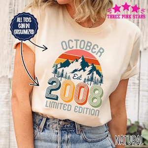 Peut inclure: Un t-shirt de couleur naturelle avec un motif de montagnes sous un coucher de soleil, incluant le texte "OCTOBER", "EST", "2008" et "LIMITED EDITION". Un badge circulaire bleu foncé à gauche indique "ALL TEXT CAN BE CUSTOMIZED".