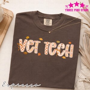 May include: A dark brown short-sleeve t-shirt with a crew neckline. The front features the text VET TECH in a light-colored, patterned font with a scalloped edge. The letters are decorated with small autumn leaves and subtle geometric designs.