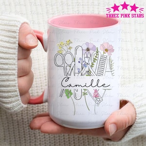 Könnte beinhalten: Weißer Keramikbecher mit rosa Innenraum und Henkel, verziert mit Blumen und Nagelwerkzeugen. Der Becher trägt den Namen "Camille" in Schreibschrift. Oben rechts steht der Text "THREE PINK STARS".
