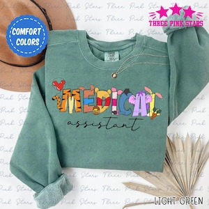 May include: Light green sweatshirt with the words "MEDICAL assistant" in colorful, cartoon-style lettering. The word "MEDICAL" features characters from a popular animated series. The sweatshirt has long sleeves and a crew neck.