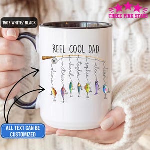Op de afbeelding: Witte keramische mok met een zwarte binnenkant en handvat. De mok heeft de tekst "REEL COOL DAD" met gepersonaliseerde namen die aan een vislijn hangen. De tekst "ALL TEXT CAN BE CUSTOMIZED" is ook zichtbaar.