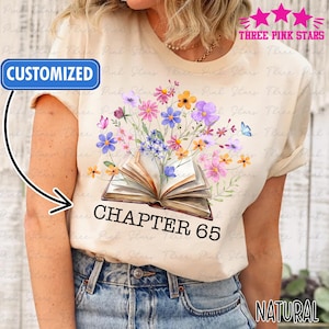 May include: Cream-colored t-shirt featuring a floral design emerging from an open book. The design includes colorful flowers and the text "CHAPTER 65". The shirt also has the words "CUSTOMIZED" and "NATURAL" printed on it.