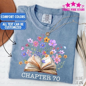 May include: Blue jean-colored t-shirt with a floral design. The graphic features an open book with colorful flowers and the text "CHAPTER 70." The shirt also has the text "COMFORT COLORS" and "ALL TEXT CAN BE CUSTOMIZED" in blue boxes.
