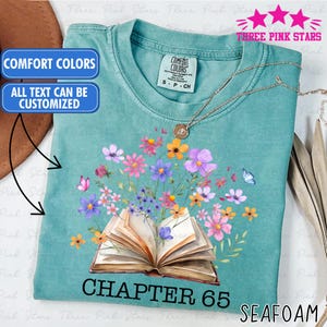 May include: Seafoam green t-shirt with a floral design. The graphic features an open book with colorful flowers and butterflies. The text "CHAPTER 65" is below the book. The shirt has a crew neck and short sleeves.