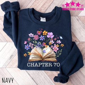 May include: Navy blue sweatshirt featuring a floral design with an open book at the center. The book is surrounded by colorful flowers and the text "CHAPTER 70". The word "NAVY" is printed at the bottom.