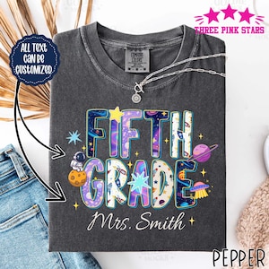 May include: A charcoal gray crew neck t-shirt featuring a space-themed graphic with the text FIFTH GRADE and Mrs. Smith. The design includes an astronaut on a planet, a purple planet with rings, a flying saucer, and various stars in vibrant colors.