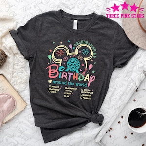 May include: A dark grey t-shirt with a colourful graphic of Mickey Mouse ears with fireworks and a globe. The text "Celebrating My Birthday Around The World" is printed below the graphic. A list of countries is printed below the text.