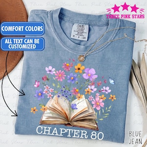 Op de afbeelding: Blauw spijkerbroekkleurig T-shirt met een bloemen- en boekafbeelding. Het ontwerp toont een open boek met kleurrijke bloemen en vlinders. De tekst "CHAPTER 80" staat op het T-shirt. Extra tekst is "COMFORT COLORS" en "ALL TEXT CAN BE CUSTOMIZED".