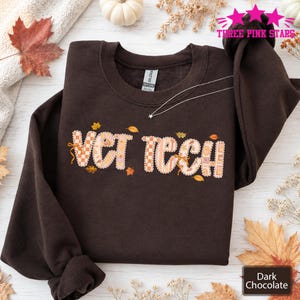 Può includere: Una felpa girocollo color marrone cioccolato scuro con la scritta VET TECH in un carattere chiaro a motivi con bordo smerlato. Le lettere sono decorate con piccole foglie autunnali e disegni a scacchi. Una sottile collana d'argento poggia sul petto.