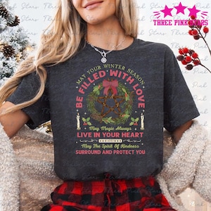 Może przedstawiać: Ciemnoszara koszulka z motywem wieńca i napisem "BE FILLED WITH LOVE". Wieniec zawiera pentagram, świece i kokardę. Koszulka zawiera również napis "May Magic Always Live in Your Heart."