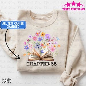 May include: Sand-colored sweatshirt featuring a floral design with an open book and the text "CHAPTER 65." The design includes colorful flowers and a butterfly. The sweatshirt has long sleeves and a crew neck.