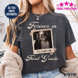 Może przedstawiać: Ciemnoszary t-shirt z krótkim rękawem z grafiką w stylu vintage polaroid, przedstawiającą czarno-białe zdjęcie dziecka w okularach przeciwsłonecznych. Napis głosi Forever in Third Grade, a na ramce polaroida widnieje Mrs. Sophia.