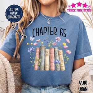 May include: A blue t-shirt featuring the text "CHAPTER 65" in black above a graphic of six vintage-style books in shades of pink, blue, and cream, surrounded by colorful watercolor-style flowers and small butterflies. The shirt has a crew neckline.