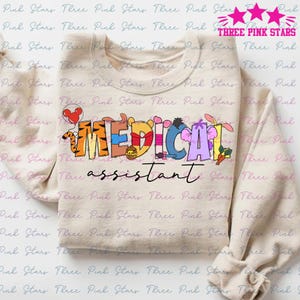 May include: A beige sweatshirt with a graphic design of Disney characters spelling out the word "MEDICAL" and the text "assistant". The characters are colorful and include Mickey Mouse, Winnie the Pooh, Tigger, and Eeyore.