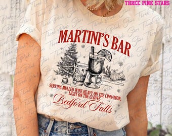 May include: Cream-colored t-shirt with a red graphic that reads "MARTINI'S BAR" above an illustration of a Christmas tree and a cocktail. The text "SERVING MULLED WINE HEAVY ON THE CINNAMON, LIGHT ON THE CLOVES" is below the illustration. The text "Bedford Falls" is at the bottom.