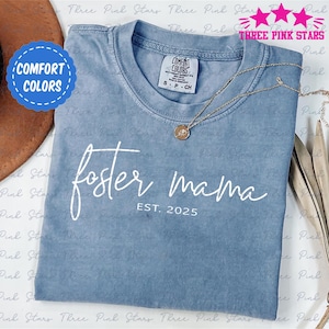 Op de afbeelding: Een lichtblauw t-shirt met de tekst "foster mama est. 2025" in wit. Het shirt is gemaakt van een zacht, comfortabel materiaal.