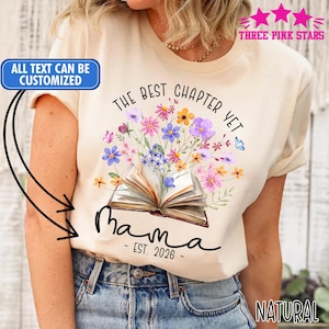 May include: Cream-colored t-shirt featuring a floral design with an open book at the center. The text "The Best Chapter Yet" and "Mama - EST. 2026" are printed on the shirt. The words "All text can be customized" are also visible.