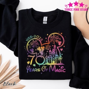May include: Black sweatshirt with a colorful graphic celebrating 70 Years of Magic. The design includes a castle, fireworks, and Mickey Mouse ears, with the years 1955-2025. The text "70 Years Of Magic" is displayed in a rainbow gradient.