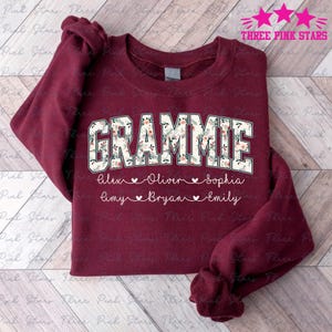 May include: A maroon sweatshirt featuring the word "GRAMMIE" in a floral design. Underneath are names: Alex, Oliver, Sophia, Amy, Bryan, and Emily. The sweatshirt has long sleeves and is a casual garment.
