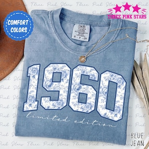 May include: Blue jean-colored t-shirt with the year "1960" in large, white, floral-patterned numbers. The shirt has the words "limited edition" printed below the numbers. A gold necklace with a pendant is draped on the shirt.