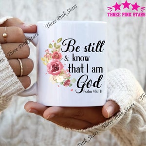 May include: White ceramic mug featuring the text "Be still & know that I am God" with Psalm 46:10 below. The mug is decorated with a floral design in shades of pink, red, and green. The mug has a white handle.
