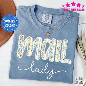 May include: A blue jean Comfort Colors t-shirt with the words "MAIL lady" in a floral design. The shirt has a crew neck and short sleeves. A gold necklace with a pendant is on the shirt.