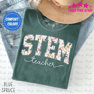 May include: A spruce green t-shirt featuring the word "STEM" in large, floral-filled letters and the word "teacher" in a cursive font. The shirt is folded on top of blue jeans, with a hat in the background. The shirt has a "Comfort Colors" tag.