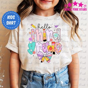 Pode incluir: Camiseta bege infantil com as palavras "hello third grade" em letras coloridas e estampadas. A camiseta apresenta um lápis, flores e um design de laço. Um círculo azul com "KIDS SHIRT" está no canto superior esquerdo.