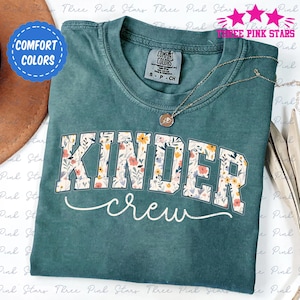 Può includere: Una maglietta Comfort Colors verde acqua con le parole "KINDER crew" in un design floreale. La maglietta ha un girocollo e maniche corte. Una collana d'argento con un ciondolo è drappeggiata sulla maglietta.