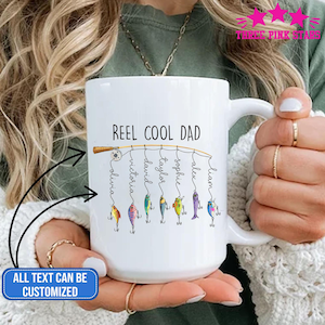 Op de afbeelding: Witte keramische mok met de tekst "REEL COOL DAD" en gepersonaliseerde namen. Het ontwerp toont een hengel met kleurrijke visbedels. De tekst "ALL TEXT CAN BE CUSTOMIZED" is ook zichtbaar.