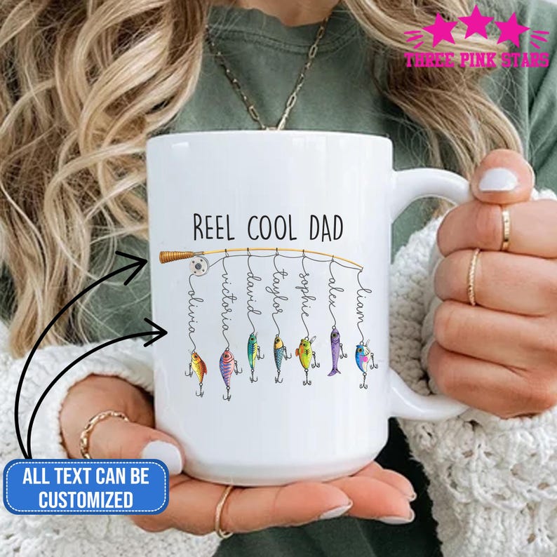 Op de afbeelding: Witte keramische mok met de tekst "REEL COOL DAD" en gepersonaliseerde namen. Het ontwerp toont een hengel met kleurrijke visbedels. De tekst "ALL TEXT CAN BE CUSTOMIZED" is ook zichtbaar.