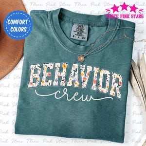 May include: Teal Comfort Colors t-shirt with "BEHAVIOR crew" in white lettering with a floral design. A gold necklace with a pendant is draped on the shirt. The shirt has a crew neck and short sleeves. A blue sticker with the words "COMFORT COLORS" is in the upper left corner.