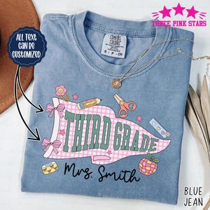 Op de afbeelding: Een blauw spijkerbroek-T-shirt met een grafisch ontwerp voor een leerkracht van de derde klas. Het ontwerp bevat de woorden "THIRD GRADE" in groen en roze, samen met schoolthema-illustraties. De tekst "Mrs. Smith" is ook opgenomen. Het shirt is van "Three Pink Stars".