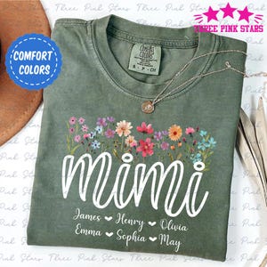 May include: A sage green t-shirt with the word "Mimi" in white script, adorned with a floral design. Below the name are the names James, Henry, Olivia, Emma, Sophia, and May. A silver necklace and a "Comfort Colors" patch are visible.
