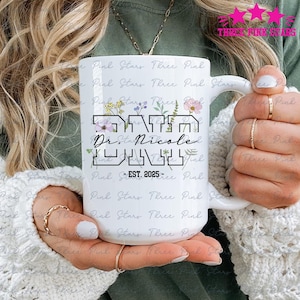 Può includere: Tazza in ceramica bianca con la scritta "DADA Dr. Nicole JUNY", decorazioni floreali e "EST. 2025". La tazza ha un manico grande. La tazza è decorata con fiori ed è tenuta da una persona.