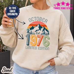 May include: A sand-colored crewneck sweatshirt featuring a graphic print with the text OCTOBER EST 1976 LIMITED EDITION. The design includes a mountain range, pine trees, and a sunset in shades of orange, blue, and green. A dark blue circular badge reads ALL TEXT CAN BE CUSTOMIZED.