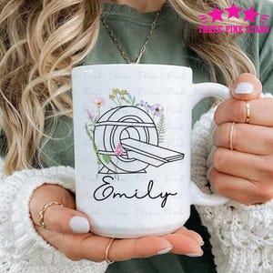 Puede incluir: Taza de cerámica blanca con una ilustración en blanco y negro de un escáner CT y detalles florales. El nombre "Emily" está escrito en cursiva. La taza tiene un asa grande.